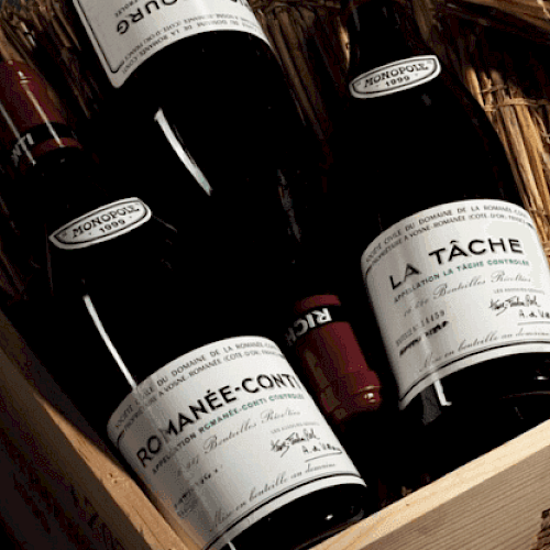 The image shows a wooden box containing several bottles of fine wine, including labels reading "ROMANÉE-CONTI" and "LA TÂCHE," nestled in straw.