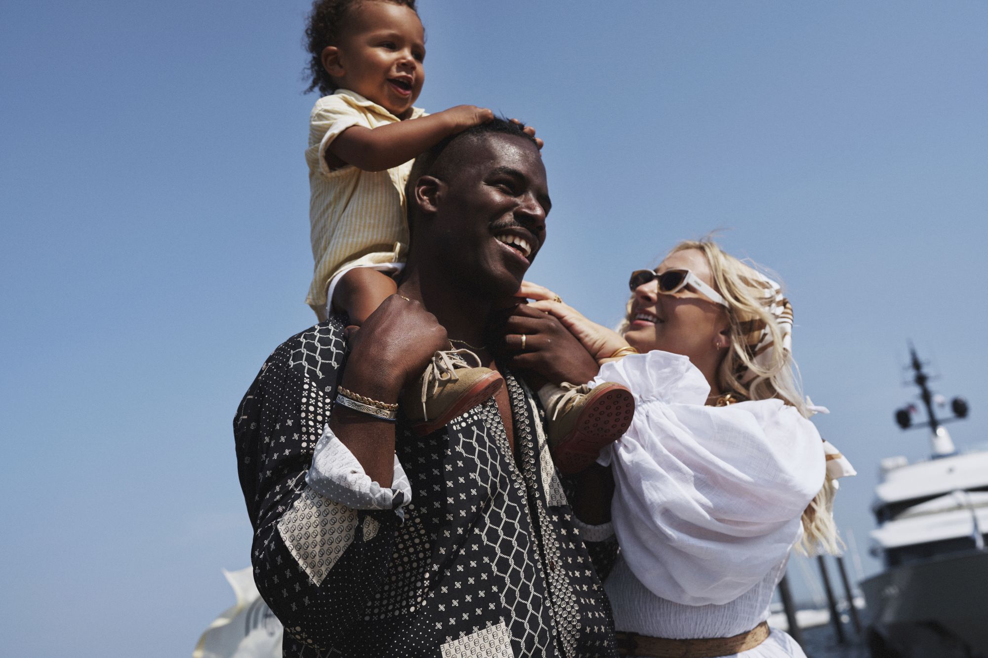 A joyful family scene: a man carries a child on his shoulders while a blonde woman hugs them, all smiling near blue sky and a boat in the background.
