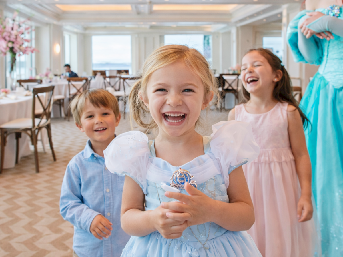 Children at a party smile and pose for a photo in a bright, elegant banquet hall with a princess in a teal dress nearby.