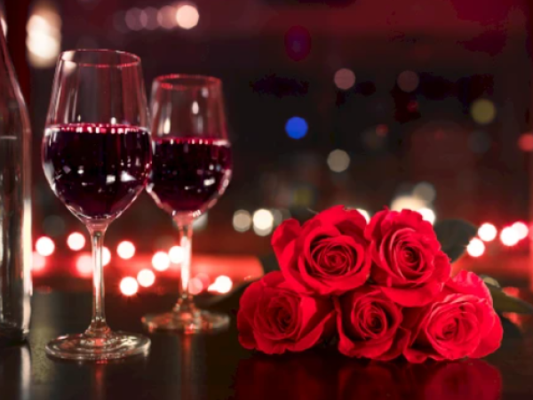 Two glasses of red wine beside a bouquet of red roses on a romantic, softly lit table. End.