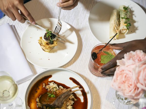 People dining in a stylish restaurant: three plates with elegant plating, a bread basket, wine glass, and a vase of pink flowers on the table.