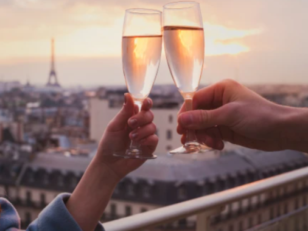 Two people toast champagne glasses against a sunset cityscape, hands warmly clinking as Paris glows in the background.
