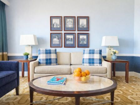 A cozy living room with a beige sofa, blue plaid cushions, two lamps, framed wall art, a round coffee table with oranges, and a rug.