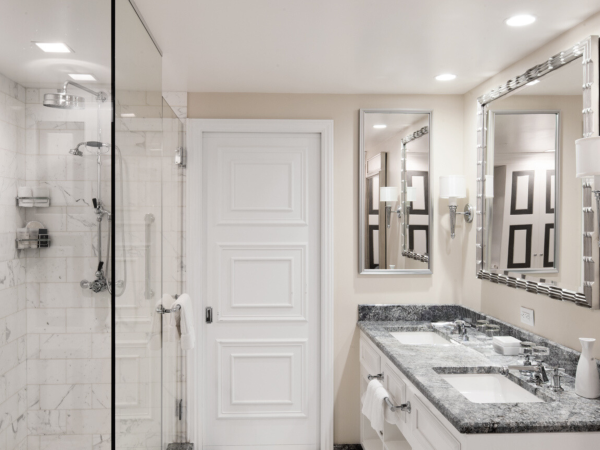 A modern bathroom with a glass-enclosed shower, white tiled walls, a double vanity with a granite countertop, two mirrors, and sleek lighting fixtures.