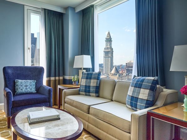 A cozy living room with a sofa, armchair, and a view of a cityscape through large windows, adorned with cushions and a lamp.