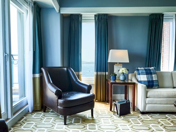 A cozy living room with a dark armchair, sofa, lamp, side table, and blue curtains in natural light.