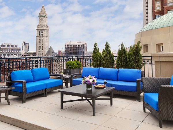 Rooftop patio with blue sofas, tables, and plants, overlooking a city.