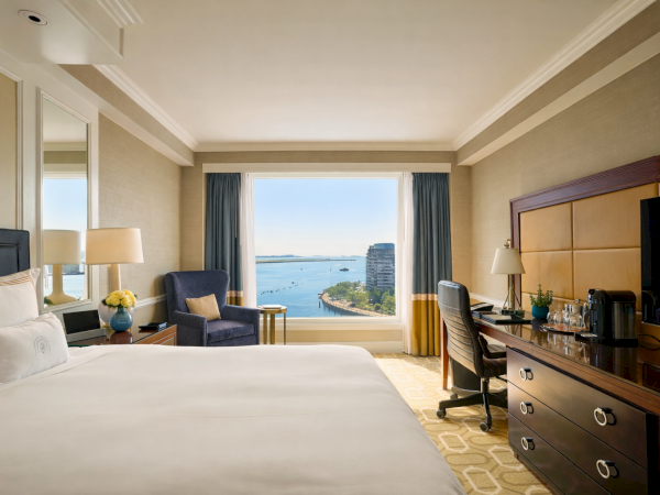 A luxurious hotel room with a large bed, two chairs, a desk, and a view of the ocean through a sunny window, with stylish lighting and d&eacute;cor.