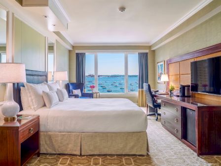 A luxury hotel bedroom with a bed, desk, TV, and large windows showing boats and water.