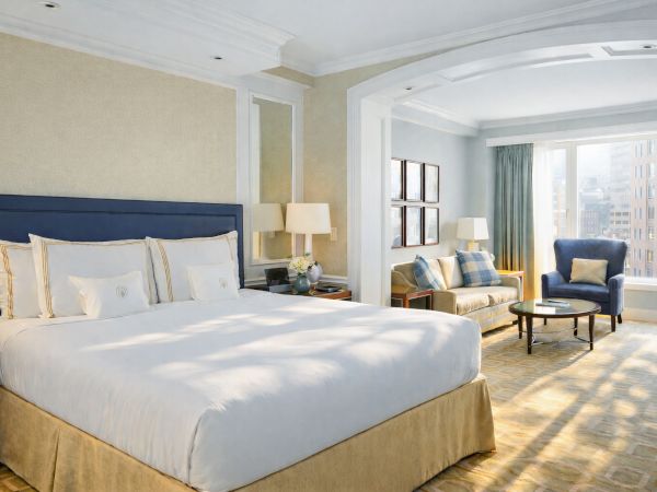 A luxurious hotel room with a king bed, beige walls, blue headboard, seating area by a window, and warm natural light streaming in.