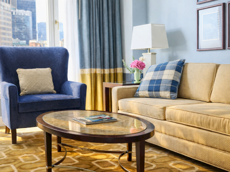 Bright hotel lounge with a blue armchair and sofa, round glass-top coffee table, lamp, flowers, curtains, and a city view.