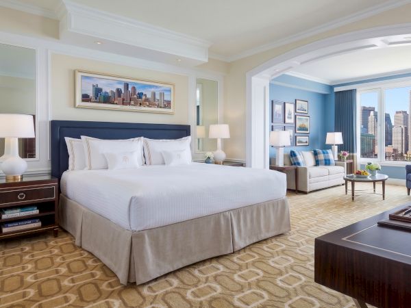 A luxurious hotel room with a king bed, blue accents, two nightstands, a sitting area, city view, and warm lighting.