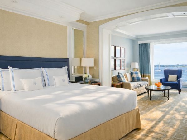 Luxurious hotel room with a king bed, beige tones, seating area, and a large window overlooking the water Coastline, inviting and bright.