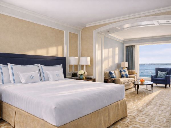 A luxurious hotel room with a large bed, beige and blue decor, a seating area by a window overlooking the ocean, and soft lighting.