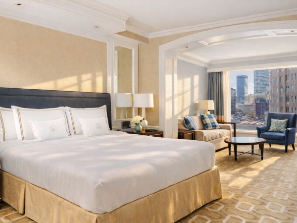 A luxurious hotel room with a plush king bed, beige decor, sitting area by a large window with city views, and warm lighting.