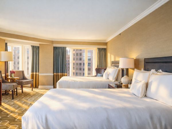 A hotel room with two large white beds, bedside lamps, a seating area by the window, warm beige walls, and soft lighting.