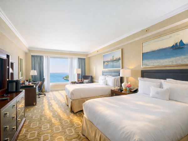 A hotel room with two large beds, a dresser, lamps, seating area, and a view of the ocean through a glass door, sunny and airy.