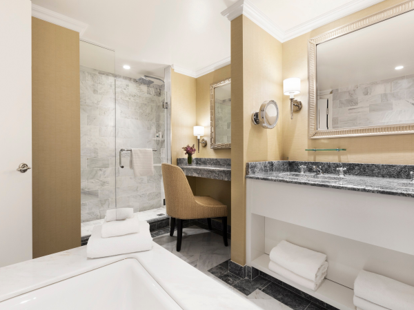 A modern hotel bathroom with a marble countertop, double sink, large mirror, glass-enclosed shower, beige walls, and a bathtub with towels.