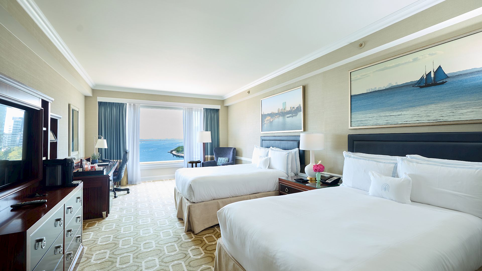 Two white beds in a bright hotel room with a sea view, a desk, and nautical wall art by the window, creating a relaxing coastal vibe.