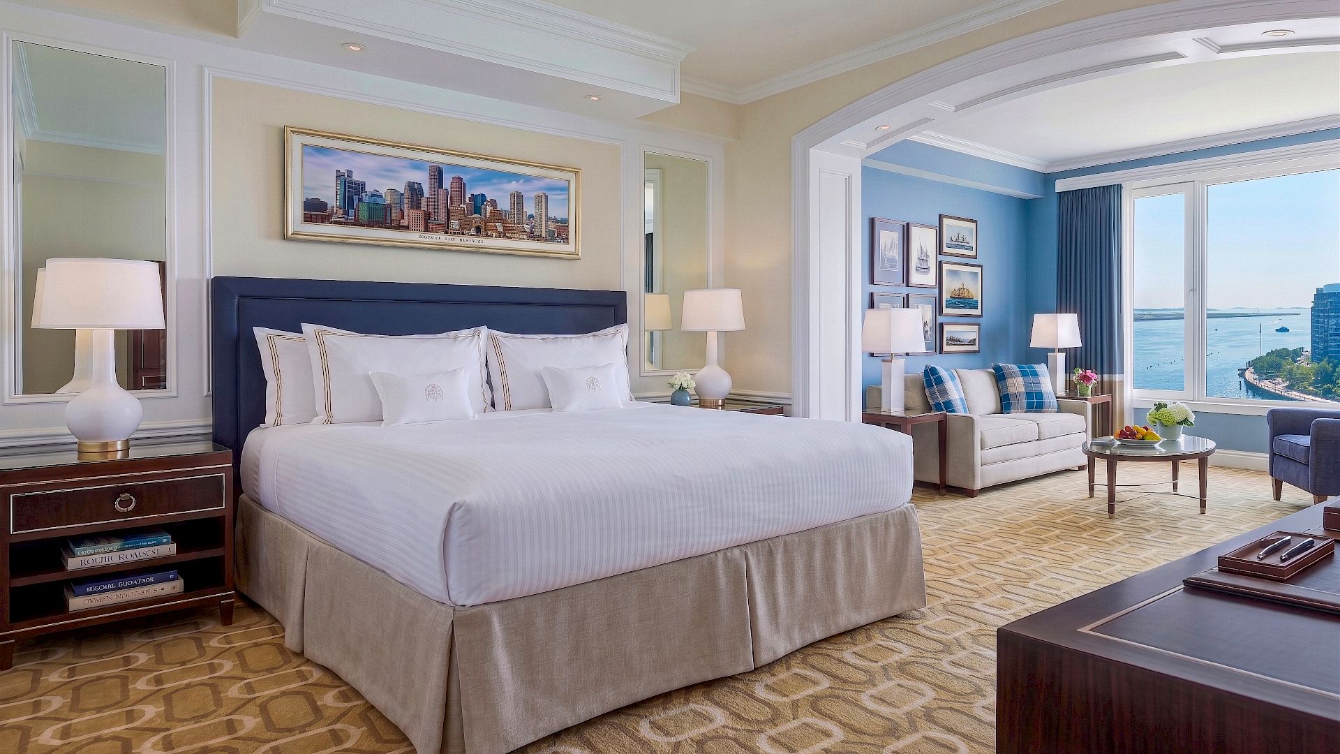 A modern hotel suite bedroom with a king bed, lamps, framed art, a seating area, and large bay windows overlooking the water.
