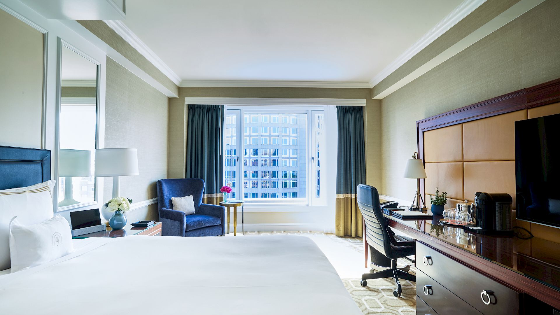 A bright hotel room with a large bed, seating area by the window, desk, and a wall-mounted TV, all in a modern, airy space.