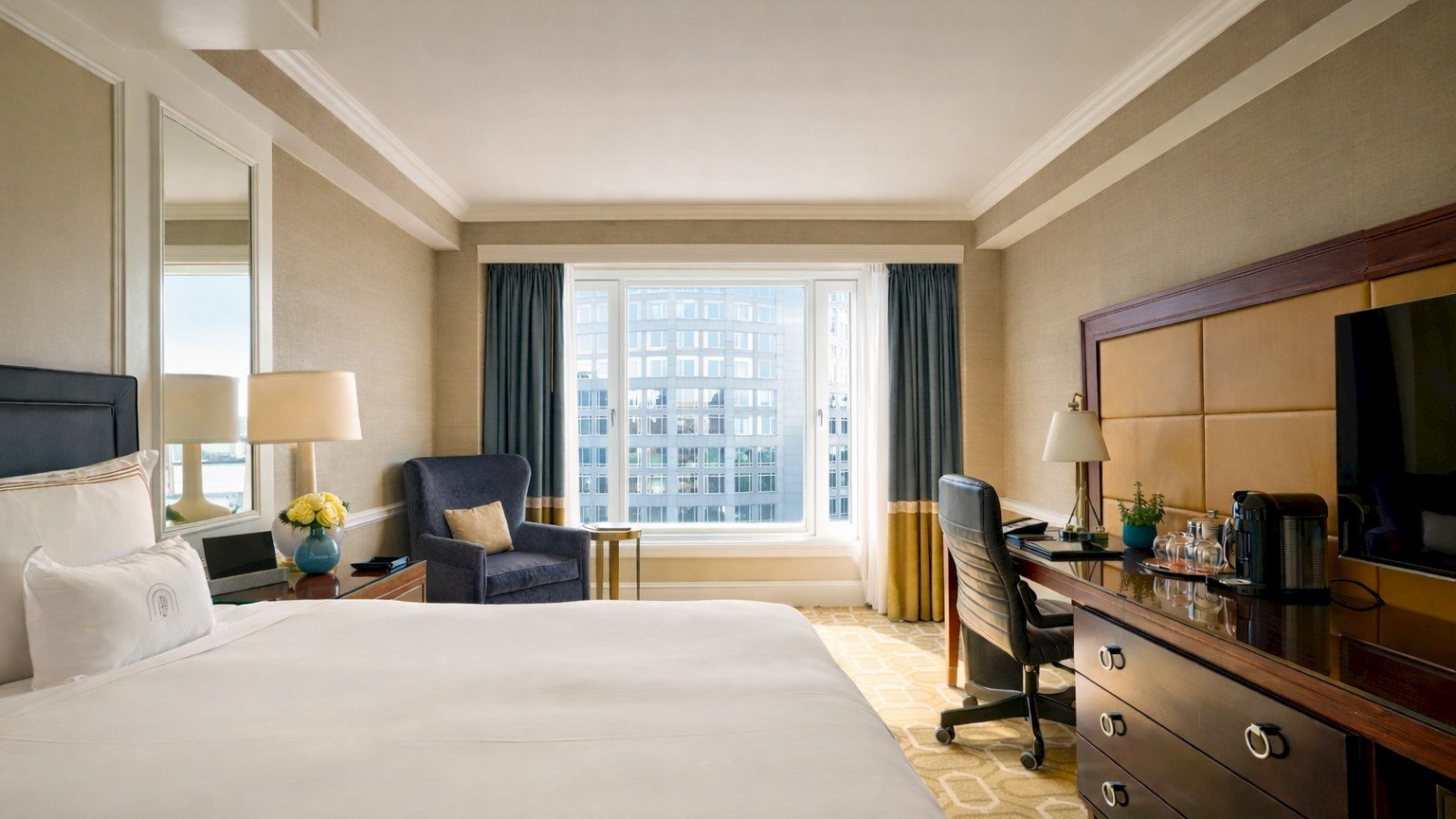 A bright hotel room with a large bed, nightstands, a seating area by the window, and a wooden dresser with a TV, all in a neutral, modern style.