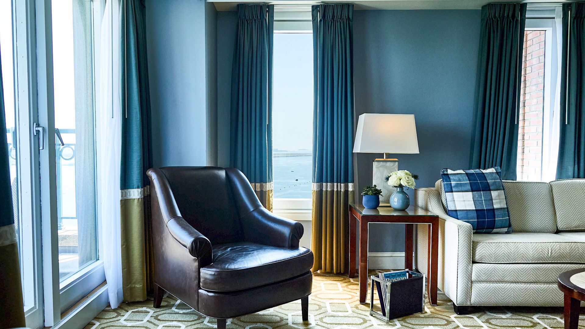 A bright hotel living room with navy walls, a black leather armchair, beige sofa, blue plaid throw, side table, lamp, and ocean view through windows.
