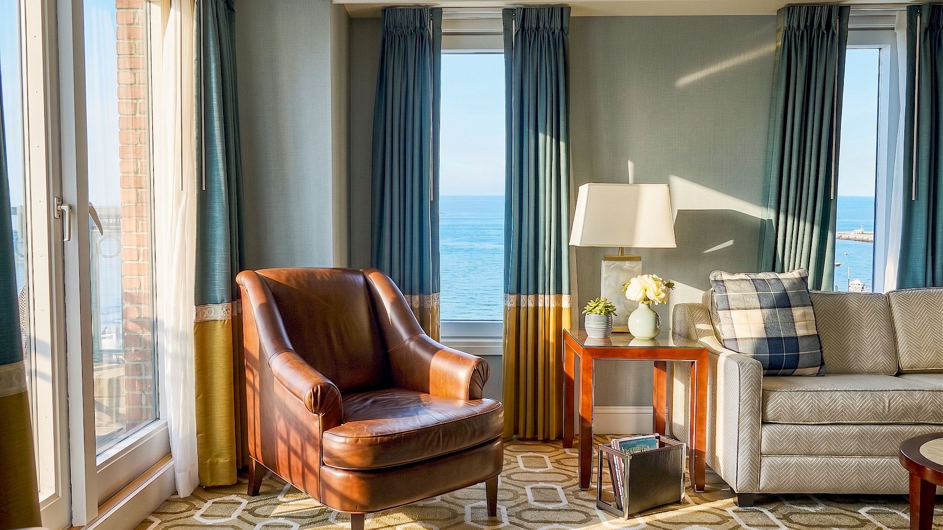 A cozy hotel lounge with leather chair, plaid sofa, small table with flowers and lamp, and teal curtains framing an ocean view.