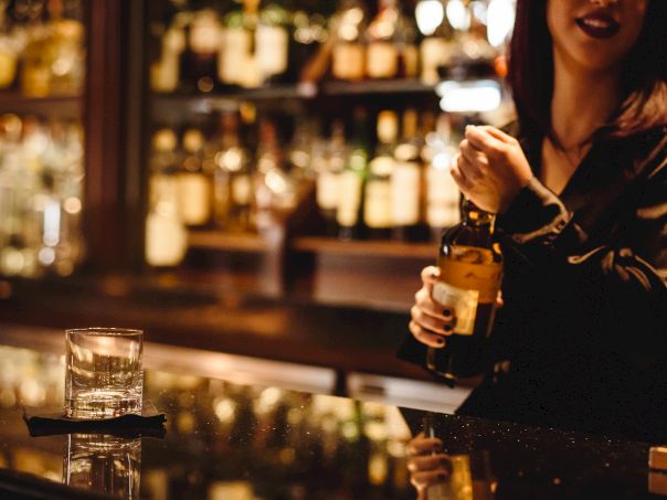 A stylish bartender pours a drink behind a glowingly lit bar; glassware, bottles, and warm ambiance reflect off the counter, creating a chic scene.