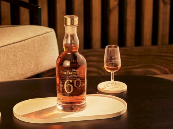 A bottle of The Balvenie 60-year single malt sits on a tray with a whiskey glass beside it, in a cozy, warm-lit setting.