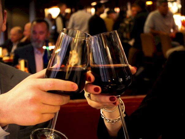Two people clink wine glasses in a dim restaurant, with bread, olives, and snacks on the table and a lively background.