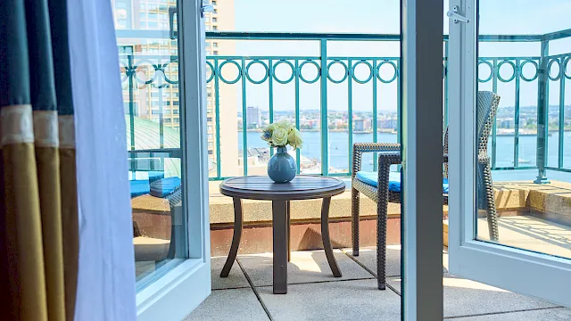 A cozy balcony scene: sliding glass doors open to a small table with a flower vase, two chairs, and a railing overlooking water and buildings.