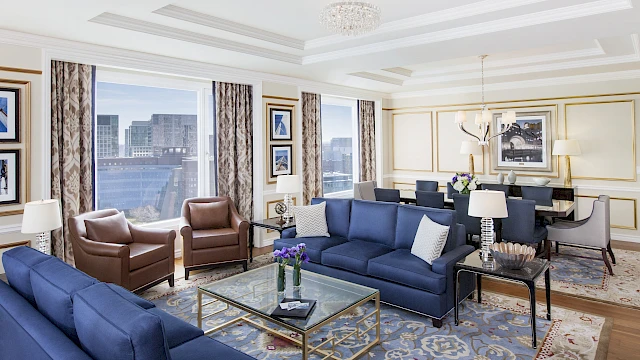 A luxurious living room with blue sofas, leather chairs, glass coffee table, patterned rug, beige walls, large windows, and elegant chandeliers.