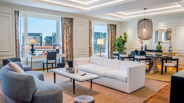 A luxurious, modern living room with white sofas, a gray lounge chair, a large area rug, and multiple desks and lamps by tall windows.