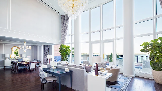 A luxurious, bright dining area with large windows, chandeliers, a blue-gray color scheme, white seating, potted plants, and a polished wooden floor.