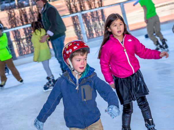 Two kids skate with others gliding on an outdoor rink; a girl in pink leads while a boy in blue follows, all in winter attire.