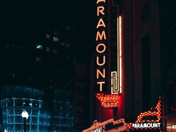 Neon-lit theater sign reads "PARAMOUNT" in a city street at night, surrounded by illuminated buildings and a dark sky.