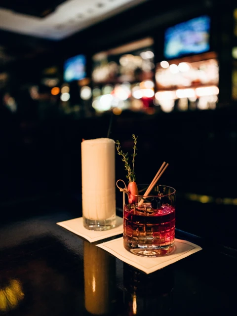 Two cocktails on a bar counter: a tall creamy drink and a short red drink with garnish and cinnamon sticks, low lighting.