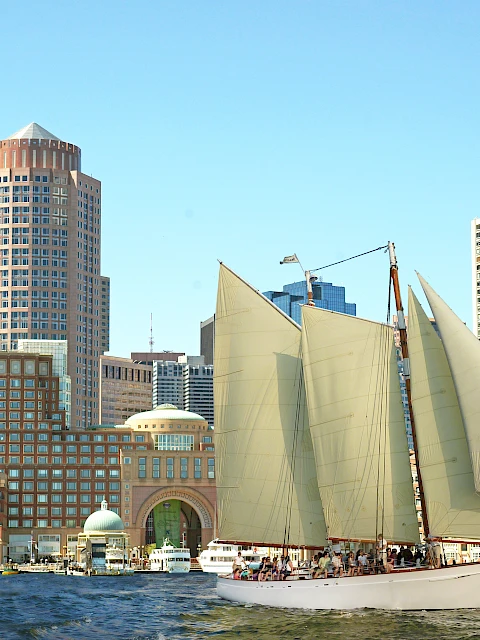 A sailboat with large cream sails glides past a city waterfront of tall buildings and a clear blue sky.