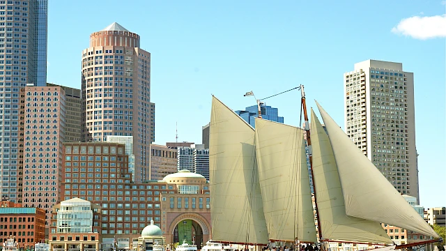 A sailboat with large cream sails glides past a city waterfront of tall buildings and a clear blue sky.