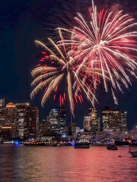 Fireworks over a city skyline at night, reflecting on the water, boats in the foreground, festive celebration lighting.