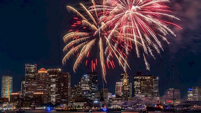 Fireworks over a city skyline at night, reflecting on the water, boats in the foreground, festive celebration lighting.