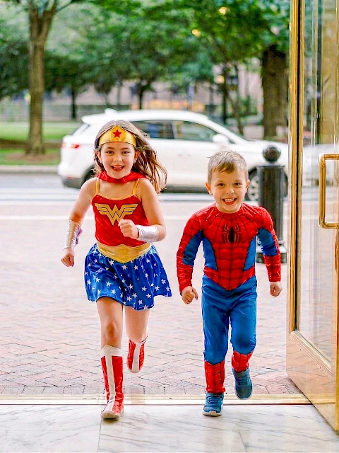 Two kids, a girl in a Wonder Woman costume and a boy in a Spider-Man suit, run hand-in-hand through open doors, smiling joyfully.