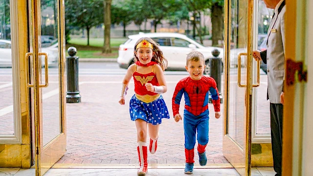 Two kids, a girl in a Wonder Woman costume and a boy in a Spider-Man suit, run hand-in-hand through open doors, smiling joyfully.