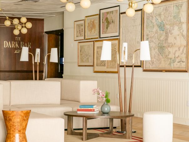 Cozy mid-century living room with white sofa, geometric rug, tall floor lamps, globe chandelier, stacked books on a coffee table, and vintage maps on the wall.