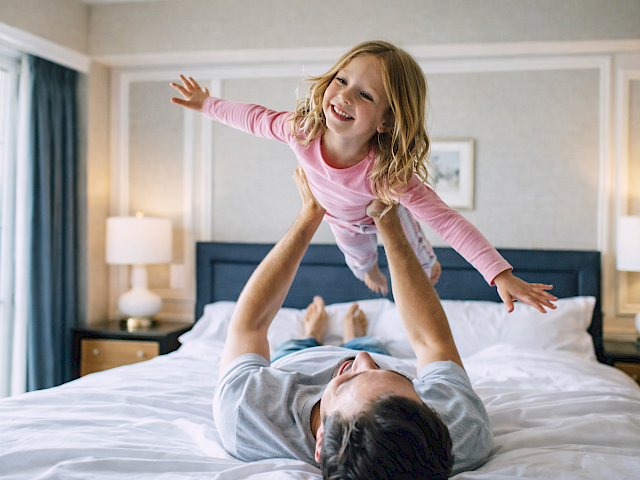 A parent lifts a smiling child in the air on a bright hotel-like bedroom with a bed, lamps, and large window, capturing a joyful moment.