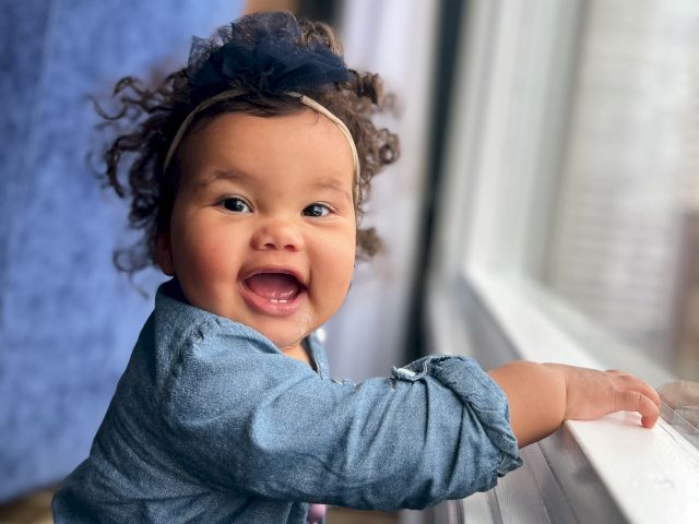A cheerful toddler with curly hair, wearing a denim dress and headband, smiles brightly while standing by a windowsill.