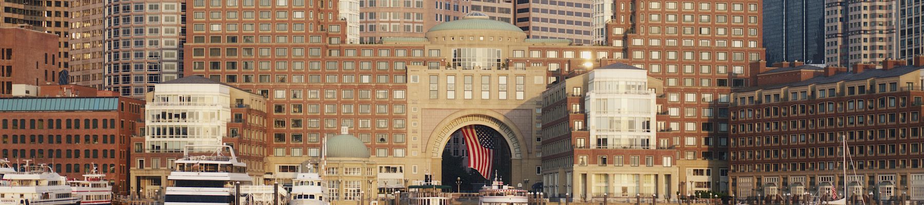 A city skyline with tall modern buildings along the waterfront, including a domed central archway and boats docked at the pier.