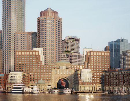 A city skyline with tall modern buildings along the waterfront, including a domed central archway and boats docked at the pier.