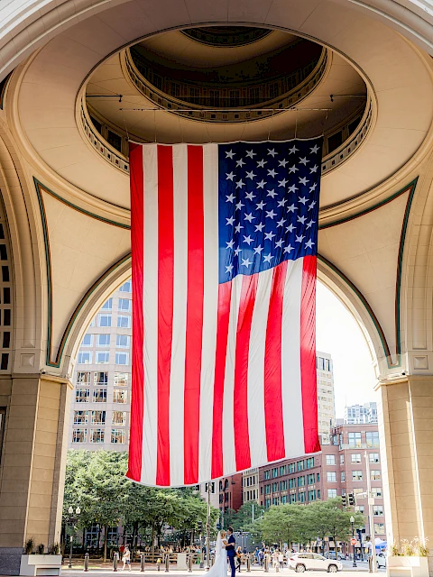A large American flag hangs between arched columns of a circular, open-air building, with a city street and trees below.